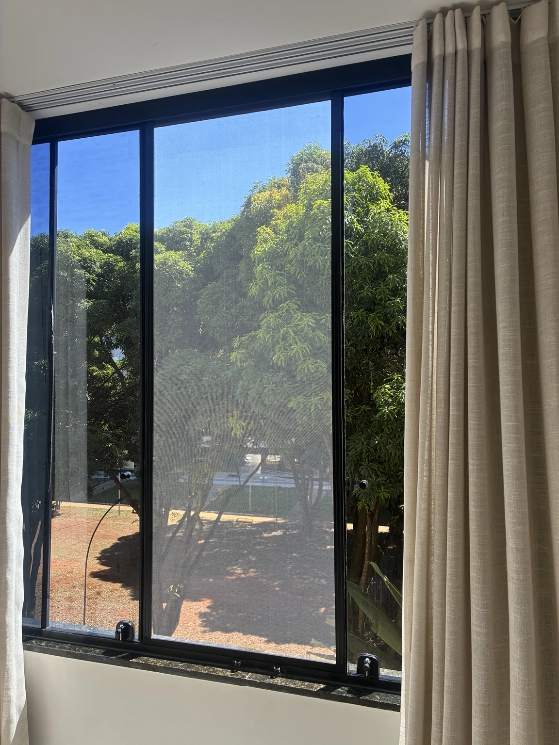 Janela ampla com tela mosquiteira entre cortinas claras e vista para as árvores na SQN 111, Bloco I, Asa Norte, Brasília DF