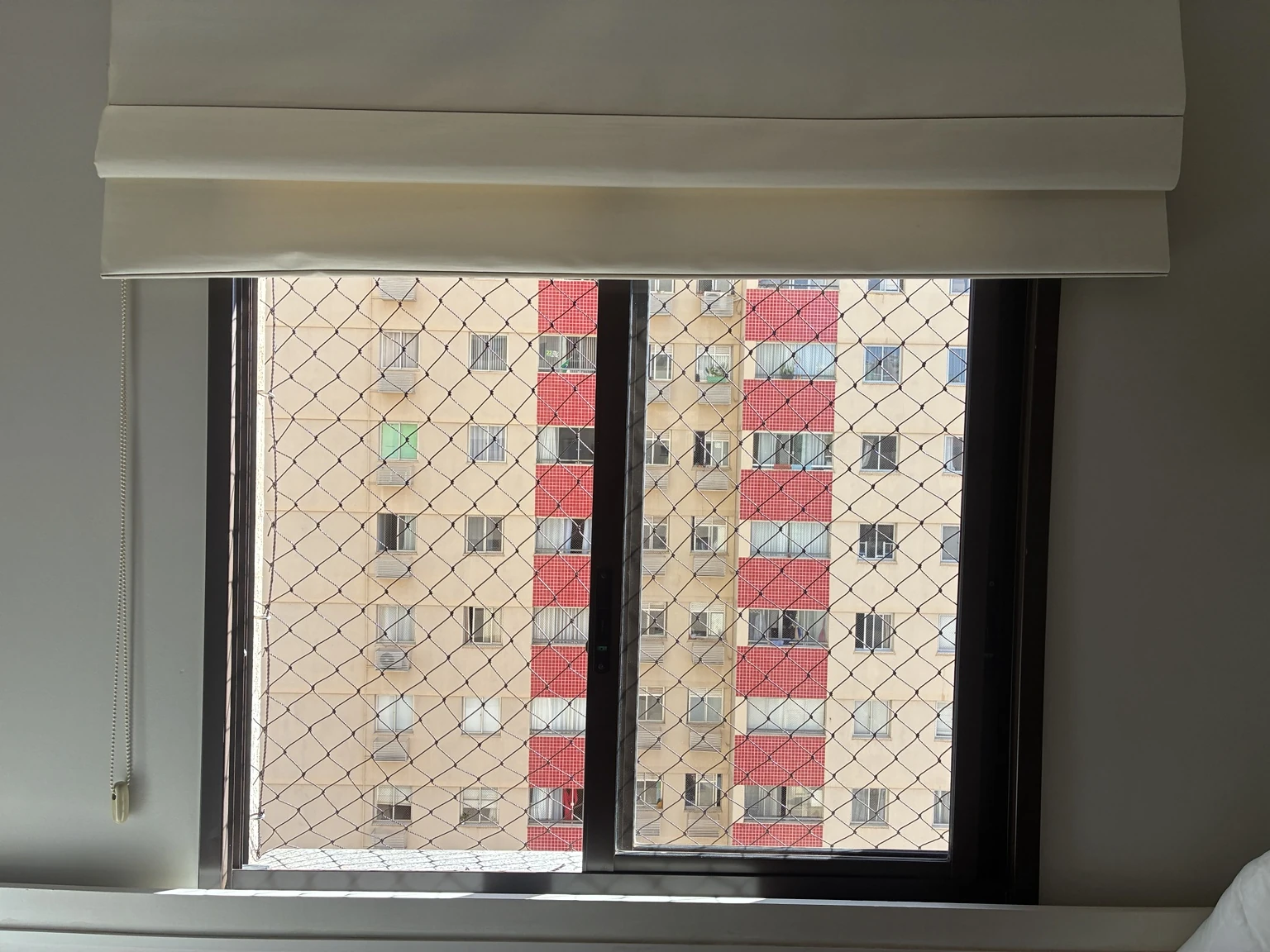 Vista frontal da janela com rede marrom e persiana no Residencial Via Naturale.