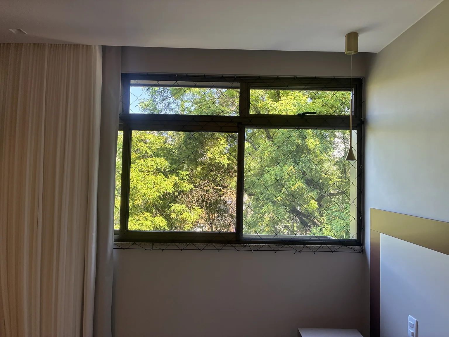 Tela mosquiteira de correr e rede preta na janela do quarto, junto às cortinas.